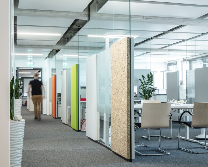 Office corridor with glass and colored acoustic panels, heating and cooling ceiling plates, view of desks and chairs, grey carpet.