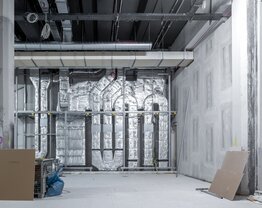 Ein großer, hoher Raum mit Betonoptik-Wänden, im Baustellenzustand. Im Hintergrund sind dick isolierte Rohre an der Wand, die miteinander verbunden sind. Schwarze Decke mit Kabelschächten.