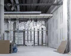 Ein großer, hoher Raum mit Betonoptik-Wänden, im Baustellenzustand. Im Hintergrund sind dick isolierte Rohre an der Wand, die miteinander verbunden sind. Schwarze Decke mit Kabelschächten.