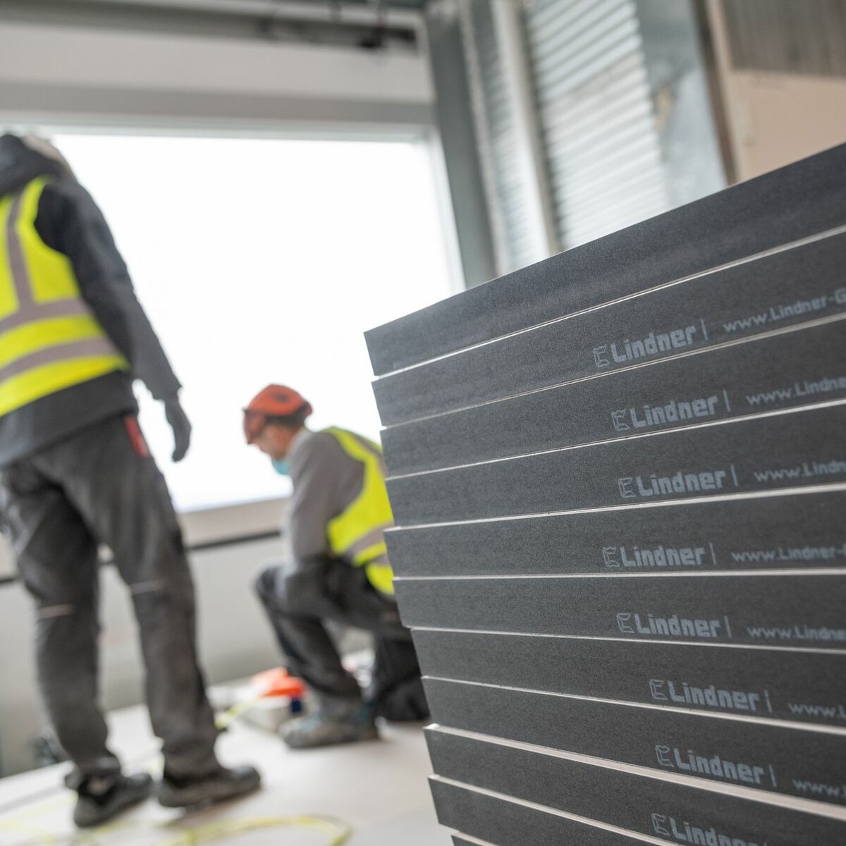 Stack of grey floor panels with Lindner company name on black side edge, blurred construction workers in neon yellow safety vests and helmets in background | © © Beat Ernst | Roche