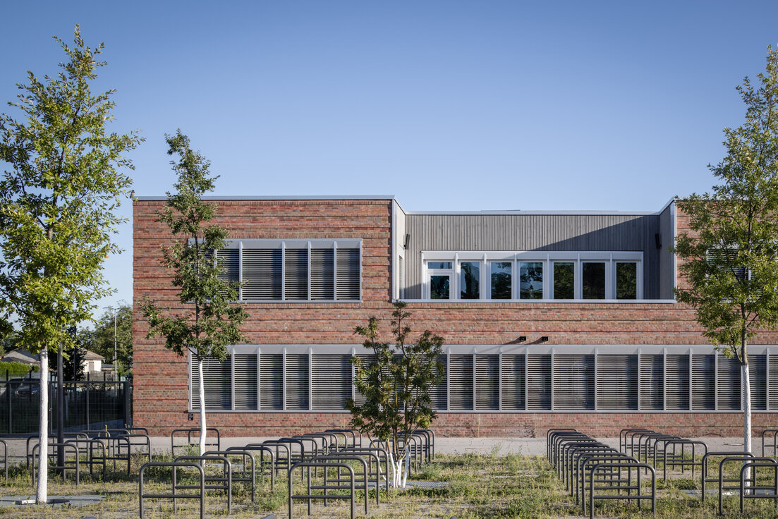 Außenansicht der Clay Schule in Berlin-Neukölln, Neubau mit rostbrauner Fassade, weißen Fenstern, dunklen Lamellen und grüner Fläche mit Bäumen im Vordergrund. | © © www.Lindner-Group.com