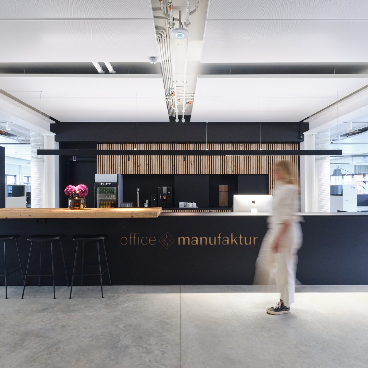 Open Work Space der Office Manufaktur in Hausham mit modernen weißen Lindner Heiz- und Kühldeckensegeln, schwarzer Teeküche mit Holzelementen und dynamischer Person im Vordergrund. | © © JANN AVERWERSER PHOTOGRAPHY