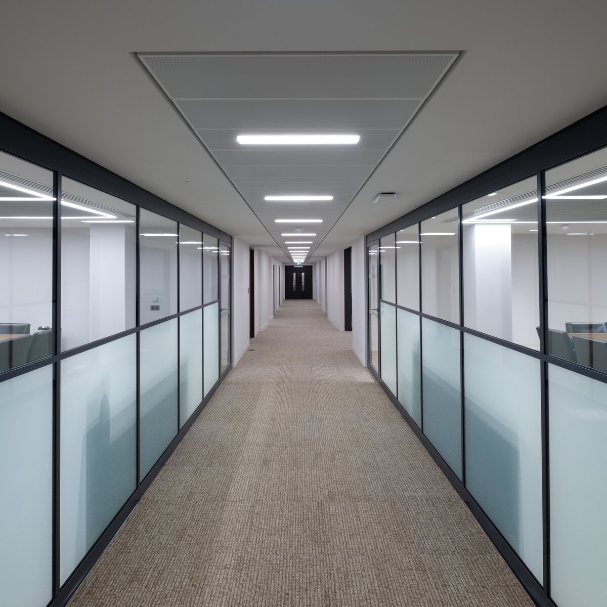 Langer Bürogebäudegang mit seitlichen Besprechungsräumen, Glaswände mit Milchglas-Folie, Holztisch, schwarze Lederstühle, brauner Teppichboden, weiße Decke mit Bandrasterdecken und integrierten Leuchten, schwarze Tür am Ende. | © © Neil Kenyon