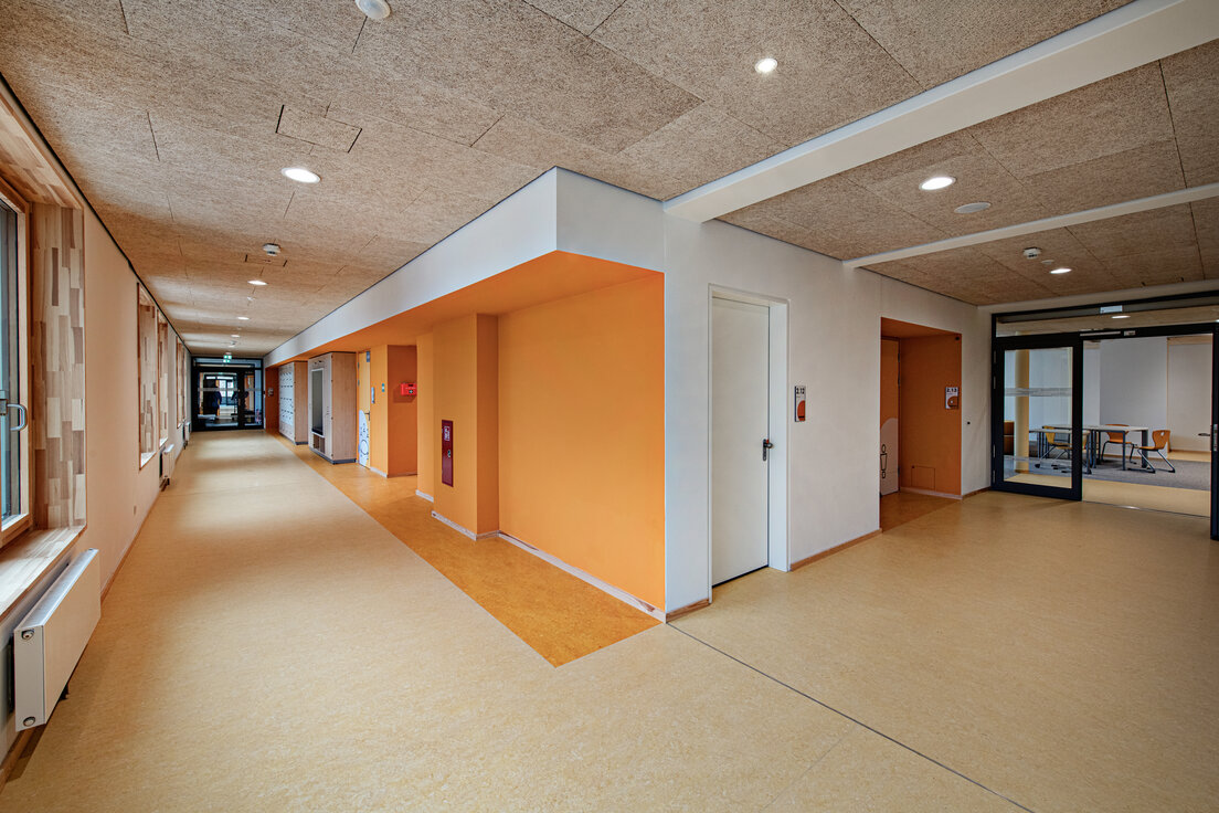 © © www.Lindner-Group.com Warmer Flurbereich der Grundschule Cottbus mit beiger Decke und orangefarbenen Wänden, Blick in Aufenthaltsraum und weiterführenden Gang | © © www.Lindner-Group.com