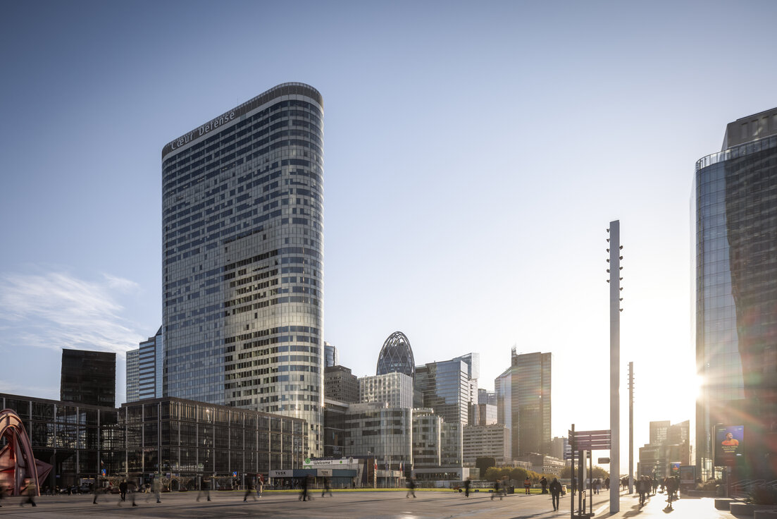 Außenansicht des Bürokomplexes Cœur Défense in Paris mit Vorplatz, umliegenden Hochhäusern, Passanten und tief stehender Sonne | © © www.Lindner-Group.com