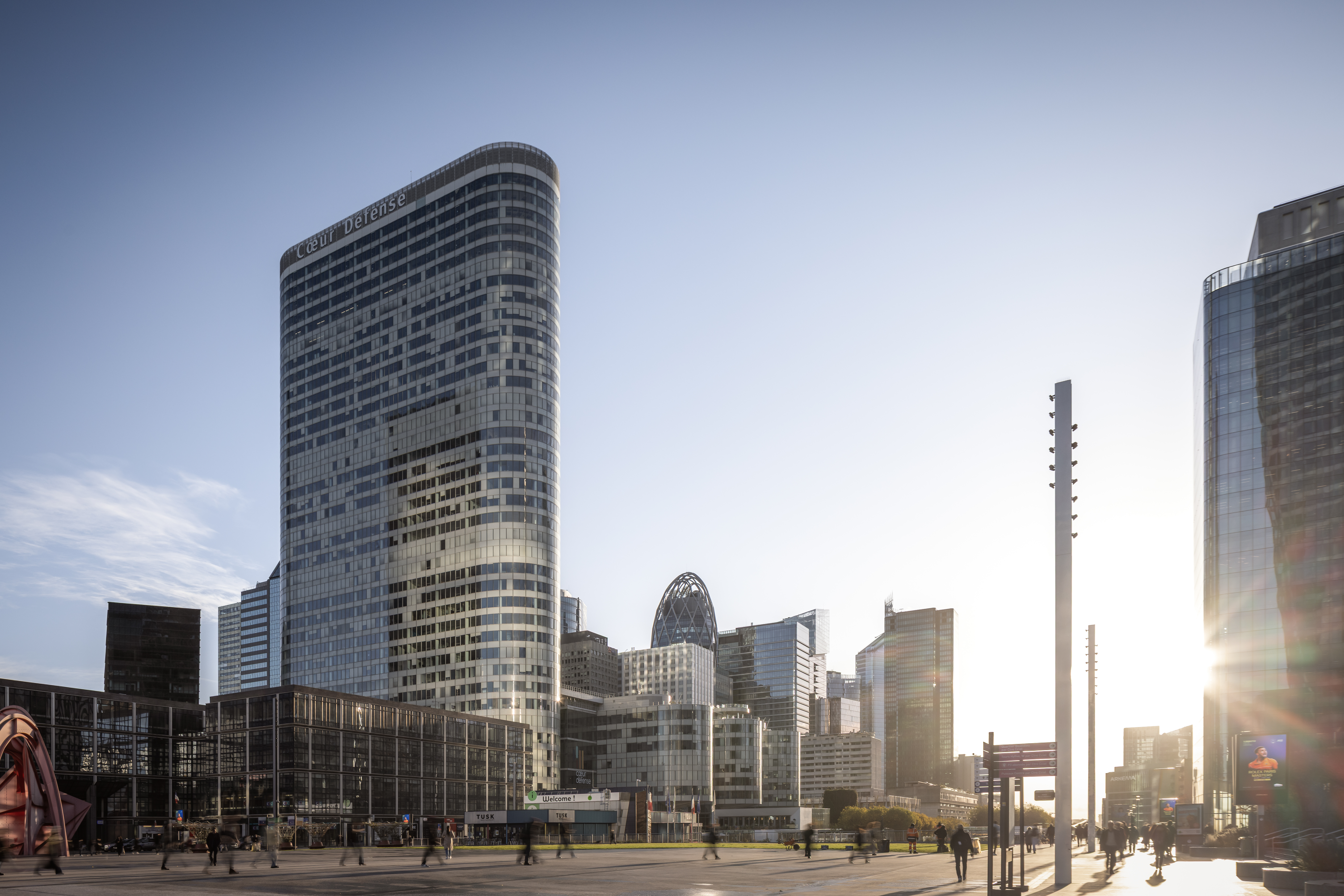 Außenansicht des Bürokomplexes Cœur Défense in Paris mit Vorplatz, umliegenden Hochhäusern, Passanten und tief stehender Sonne | © © www.Lindner-Group.com